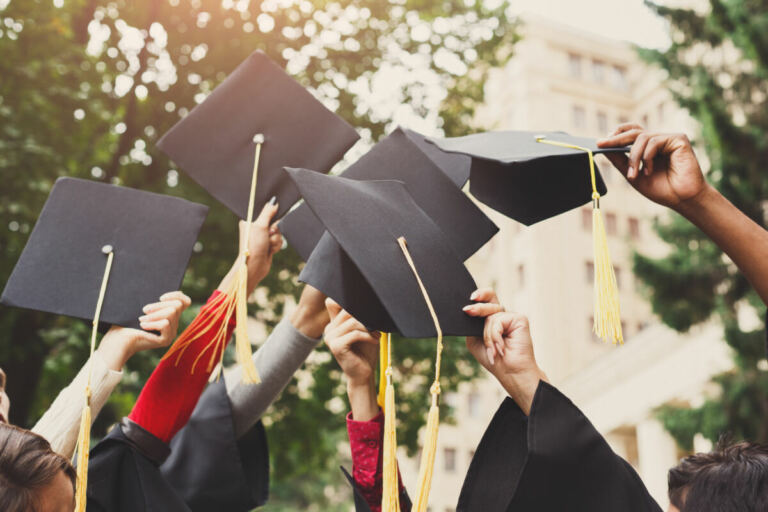 Several hands hold up black graduation caps with yellow tassels