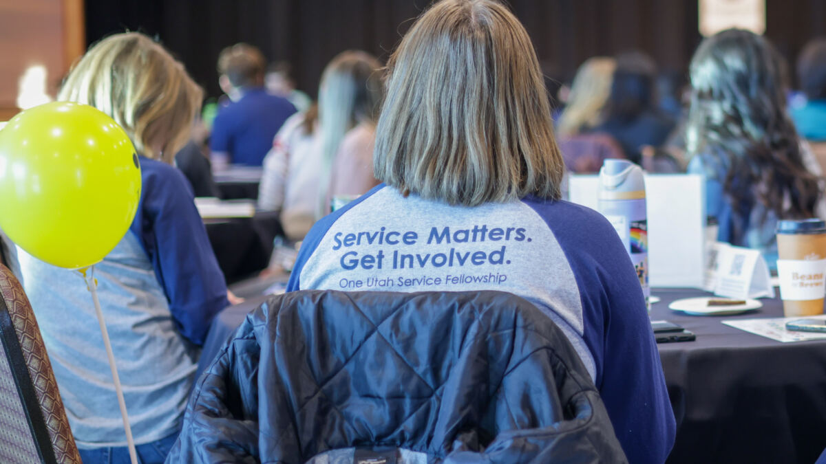 Attendees at a conference listen to a speaker; a participant in the foreground wears a shirt reading, “Service Matters. Get Involved.”