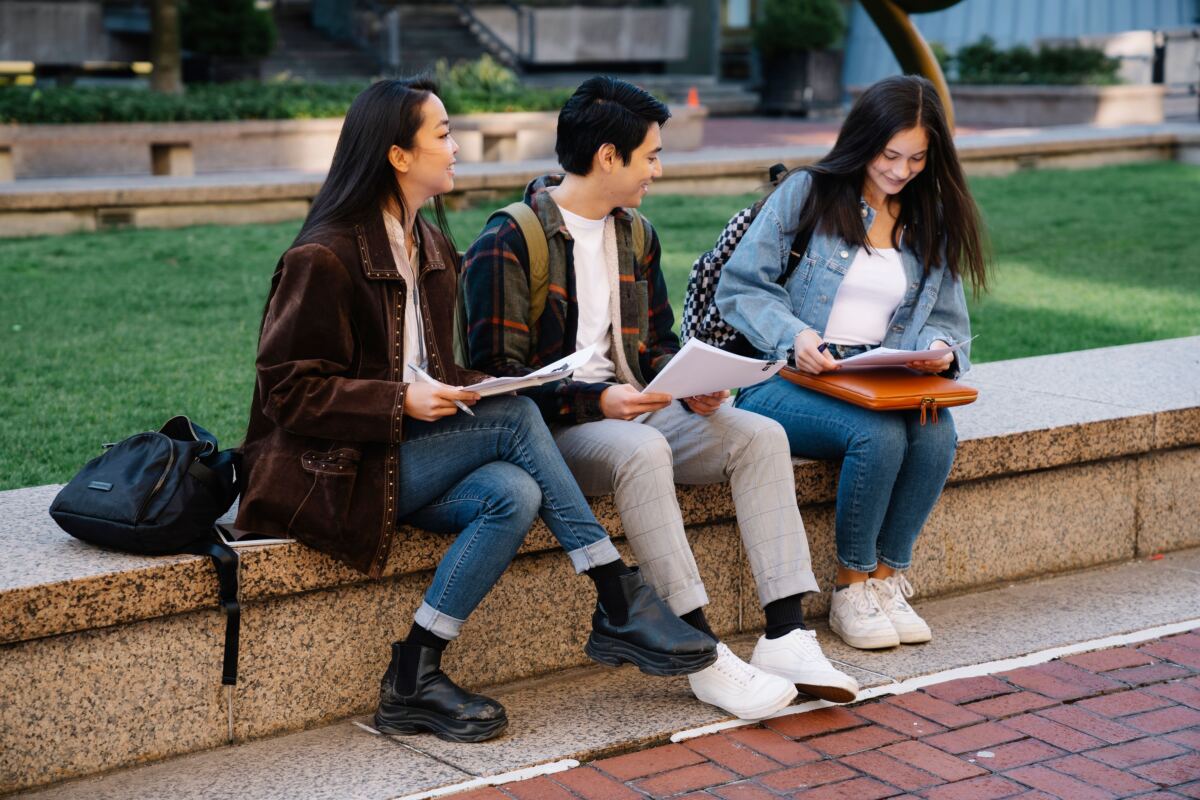 Three college students sitting outside with papers in hand.